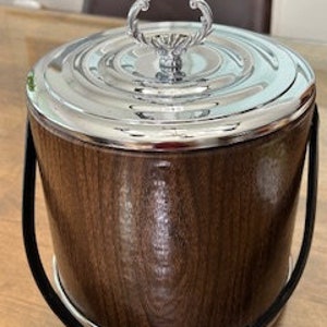 May include: A brown wooden ice bucket with a chrome lid and handle. The bucket has a black leather carrying strap.
