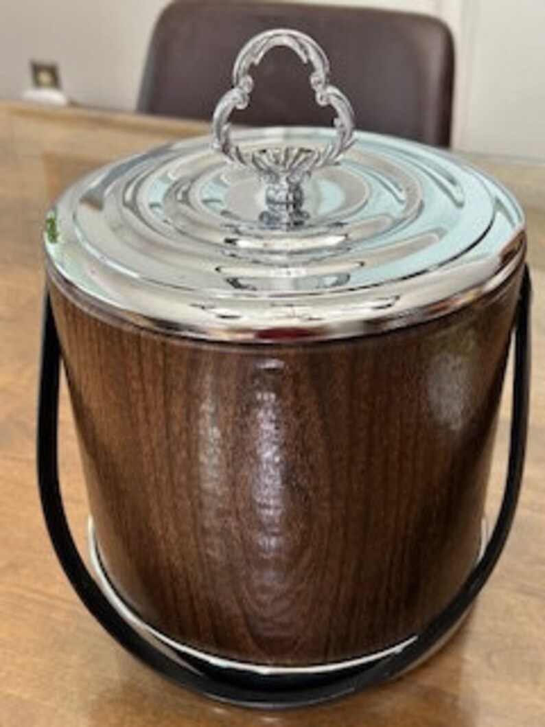 May include: A brown wooden ice bucket with a chrome lid and handle. The bucket has a black leather carrying strap.