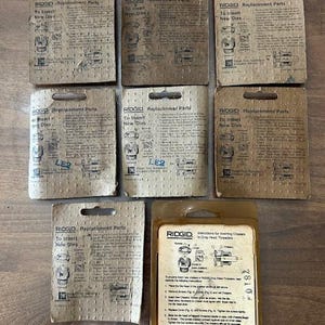 May include: A collection of RIDGID replacement parts packages. The packages are various shades of brown and tan, with text and diagrams. One package is in a clear plastic casing, displaying instructions for inserting chasers in drop head threaders.