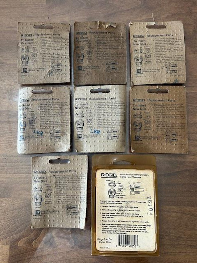 May include: A collection of RIDGID replacement parts packages. The packages are various shades of brown and tan, with text and diagrams. One package is in a clear plastic casing, displaying instructions for inserting chasers in drop head threaders.