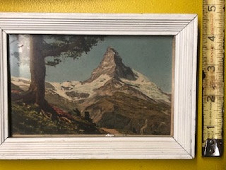 Peut inclure: Une estampe vintage encadr&eacute;e repr&eacute;sentant un paysage de montagne avec un sommet enneig&eacute;, un arbre vert et un premier plan rocheux.