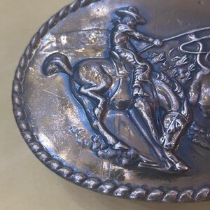 May include: Oval silver-toned belt buckle featuring a cowboy on a bucking bronco, roping a steer. The detailed relief shows the cowboy in a hat, with a rope in hand. The buckle has a rope-like border.