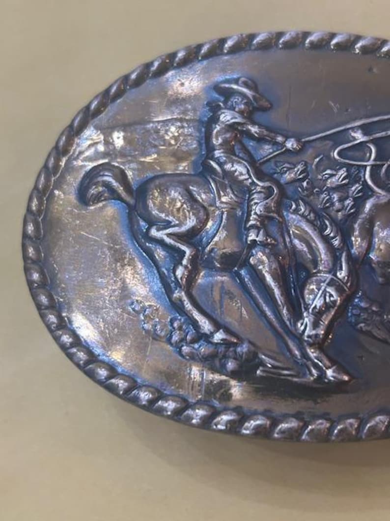 May include: Oval silver-toned belt buckle featuring a cowboy on a bucking bronco, roping a steer. The detailed relief shows the cowboy in a hat, with a rope in hand. The buckle has a rope-like border.