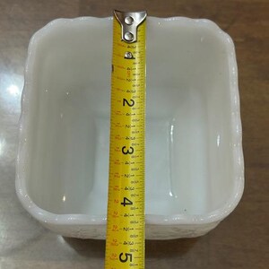 May include: A square white milk glass bowl with a scalloped rim and embossed floral detailing on the exterior. A yellow measuring tape is placed across the center, showing the bowl has a width of approximately 5.5 inches.