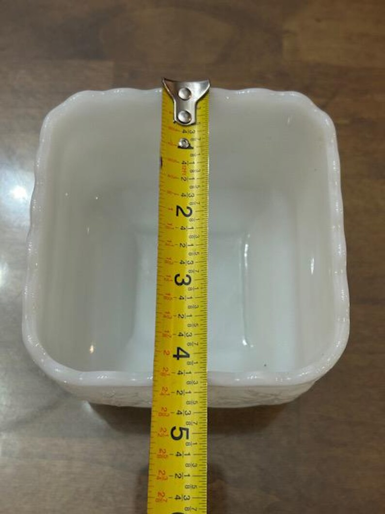 May include: A square white milk glass bowl with a scalloped rim and embossed floral detailing on the exterior. A yellow measuring tape is placed across the center, showing the bowl has a width of approximately 5.5 inches.