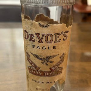 May include: A vintage glass jar with a metal lid. The jar has a label with the text "De Voe's Eagle Pure Extra Quality Snuff". The label features an eagle illustration. The glass is clear, and the label is aged.