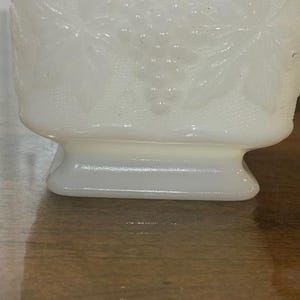 May include: A white milk glass vase featuring a raised, embossed pattern of grape clusters and leaves. The surface has a textured, stippled background detail. The vase has a square-shaped body that tapers down to a solid, smooth pedestal base.