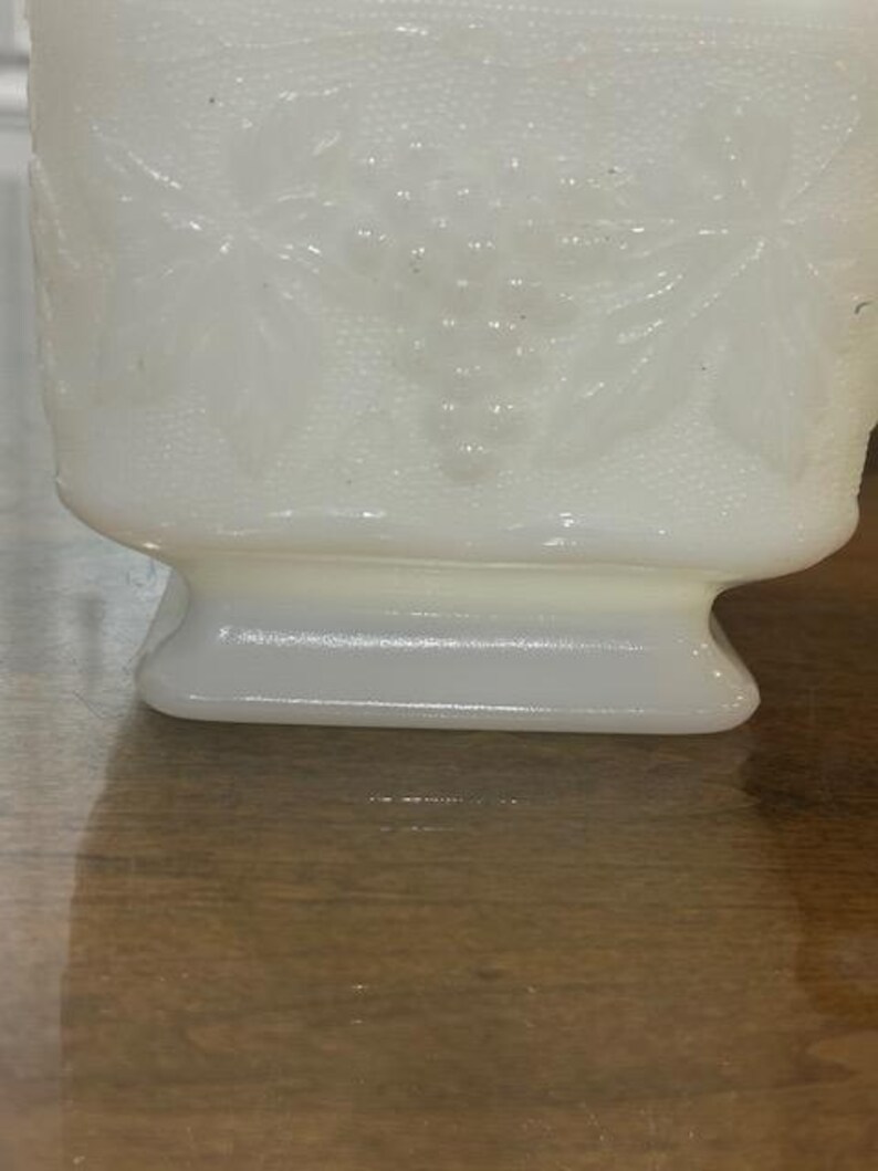 May include: A white milk glass vase featuring a raised, embossed pattern of grape clusters and leaves. The surface has a textured, stippled background detail. The vase has a square-shaped body that tapers down to a solid, smooth pedestal base.