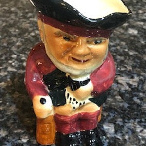 May include: A ceramic figurine of a man in a tricorn hat and a red coat, holding a cane. The figurine has a jovial expression and is painted with detailed features. The base is black and the figurine is approximately 4 inches tall.