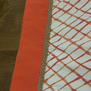 Puede incluir: Una pieza textil con un panel vertical naranja s&oacute;lido a la izquierda, separado por un borde trenzado de yute color canela de una secci&oacute;n blanca a la derecha. La secci&oacute;n blanca muestra un patr&oacute;n de cuadr&iacute;cula de l&iacute;neas naranjas y rojas.