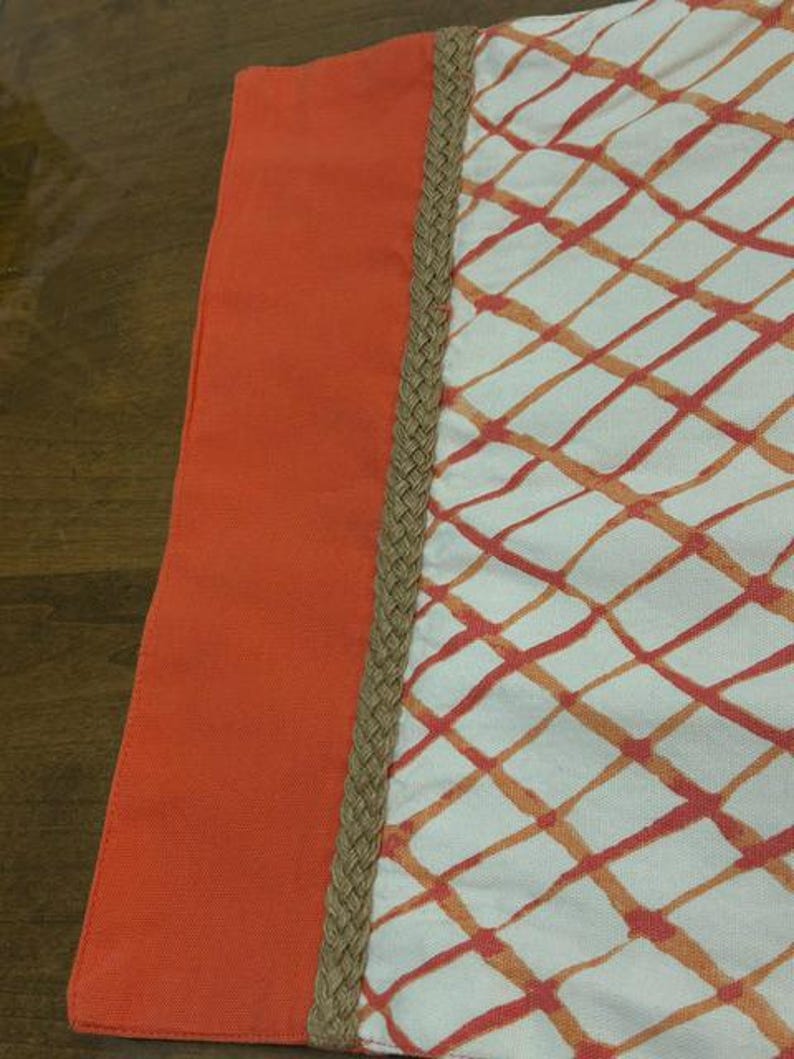 Puede incluir: Una pieza textil con un panel vertical naranja s&oacute;lido a la izquierda, separado por un borde trenzado de yute color canela de una secci&oacute;n blanca a la derecha. La secci&oacute;n blanca muestra un patr&oacute;n de cuadr&iacute;cula de l&iacute;neas naranjas y rojas.
