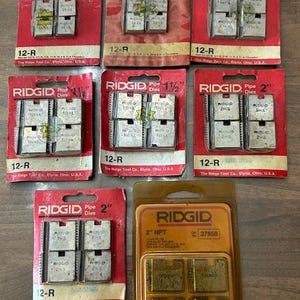 May include: A collection of RIDGID pipe dies in various sizes, including 1/2", 3/4", 1", 1 1/2", and 2". The dies are packaged in red and orange cardboard with the RIDGID logo and size information. The 2" NPT dies are in a clear plastic package.