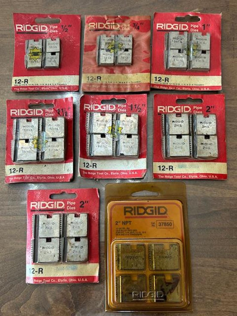 May include: A collection of RIDGID pipe dies in various sizes, including 1/2", 3/4", 1", 1 1/2", and 2". The dies are packaged in red and orange cardboard with the RIDGID logo and size information. The 2" NPT dies are in a clear plastic package.