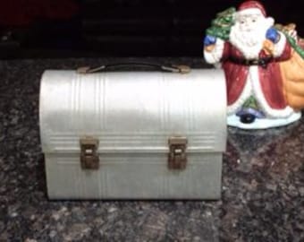 May include: A vintage silver metal lunchbox with a black handle and two latches. The lunchbox has a ribbed design.