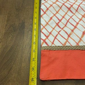 Puede incluir: Un camino de mesa rectangular con fondo blanco y un patr&oacute;n de cuadr&iacute;cula diagonal en naranja y rojo. Un borde trenzado color canela separa la secci&oacute;n estampada de una franja naranja s&oacute;lida. Una cinta m&eacute;trica amarilla indica 40,6 cm.