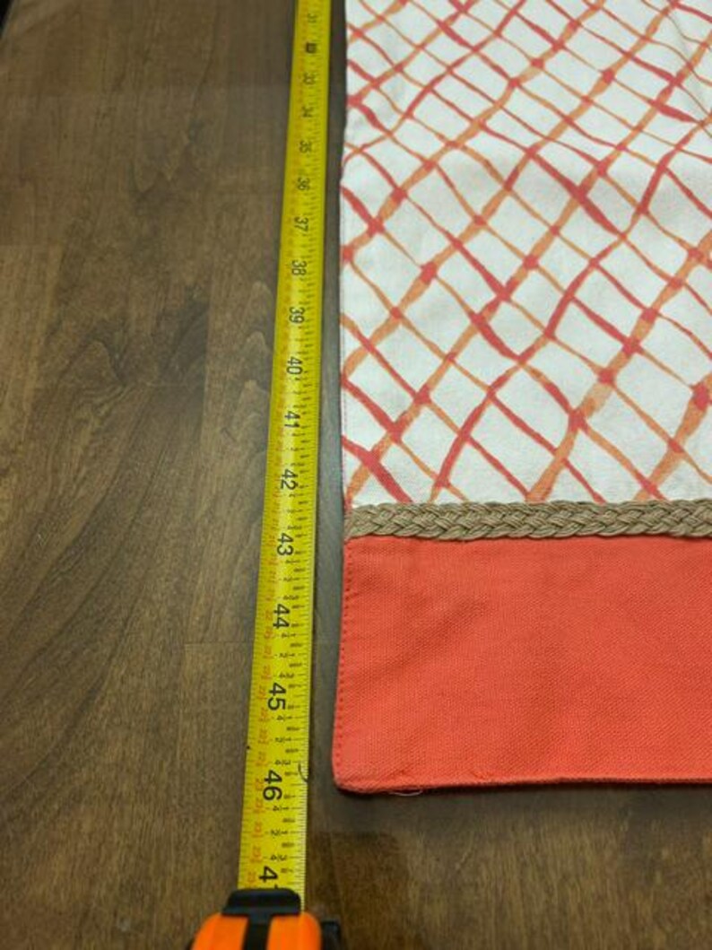 Puede incluir: Un camino de mesa rectangular con fondo blanco y un patr&oacute;n de cuadr&iacute;cula diagonal en naranja y rojo. Un borde trenzado color canela separa la secci&oacute;n estampada de una franja naranja s&oacute;lida. Una cinta m&eacute;trica amarilla indica 40,6 cm.