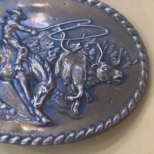 May include: Oval-shaped, silver-toned belt buckle with a detailed relief of a cowboy roping a bull. The buckle has a rope-like border and depicts a cowboy on horseback, lassoing a bull in a dynamic scene. A classic Western-themed accessory.