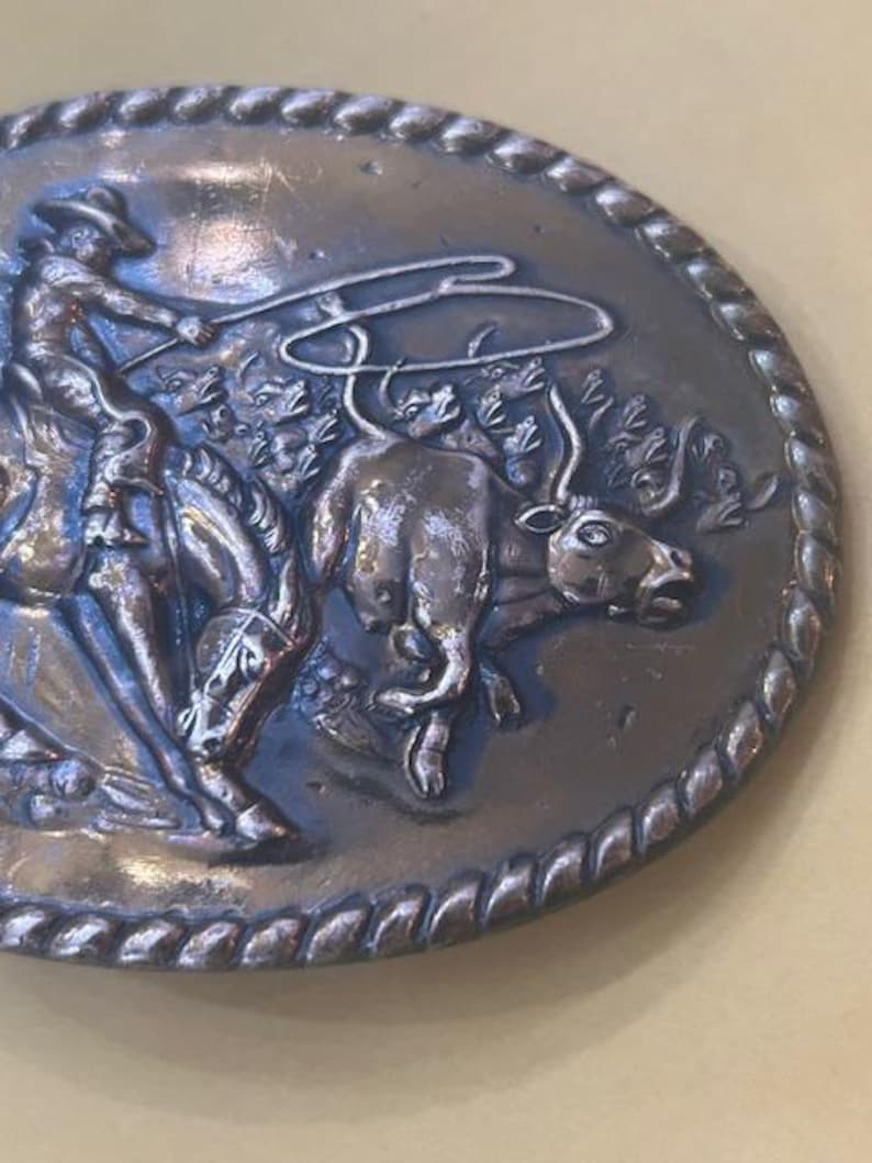 May include: Oval-shaped, silver-toned belt buckle with a detailed relief of a cowboy roping a bull. The buckle has a rope-like border and depicts a cowboy on horseback, lassoing a bull in a dynamic scene. A classic Western-themed accessory.