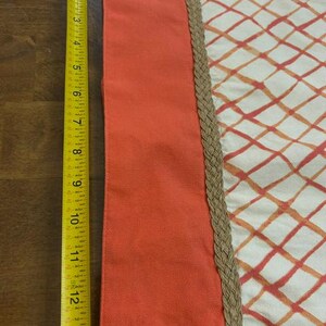Puede incluir: Un art&iacute;culo de tela rectangular con un panel naranja s&oacute;lido a la izquierda y una secci&oacute;n color crema con un patr&oacute;n de cuadr&iacute;cula de diamantes naranja y rojo a la derecha, separados por un borde trenzado color canela. Una cinta m&eacute;trica amarilla mide 33 cm.