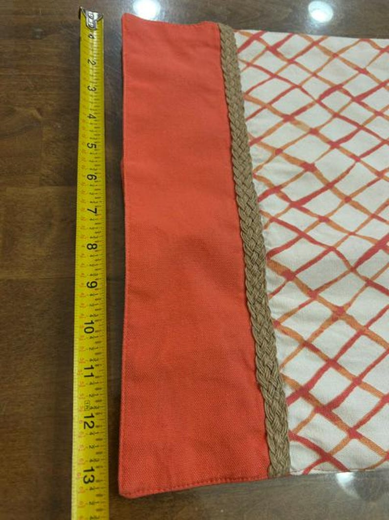 Puede incluir: Un art&iacute;culo de tela rectangular con un panel naranja s&oacute;lido a la izquierda y una secci&oacute;n color crema con un patr&oacute;n de cuadr&iacute;cula de diamantes naranja y rojo a la derecha, separados por un borde trenzado color canela. Una cinta m&eacute;trica amarilla mide 33 cm.