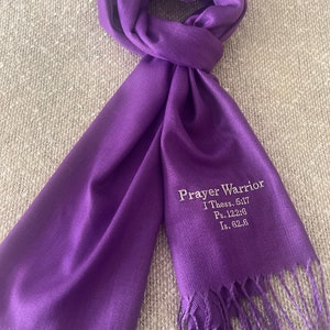 May include: A vibrant purple scarf with a fringed edge. The scarf features the embroidered text "Prayer Warrior" along with Bible verses. The scarf is made of a soft material and is draped on a neutral surface.