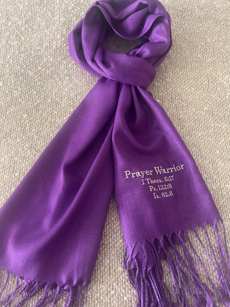 May include: A vibrant purple scarf with a fringed edge. The scarf features the embroidered text "Prayer Warrior" along with Bible verses. The scarf is made of a soft material and is draped on a neutral surface.