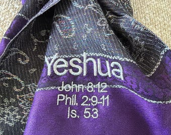 May include: A purple scarf with the embroidered word "Yeshua" in white. Below the word are the references John 8:12, Phil. 2:9-11, and Is. 53. The scarf has a patterned design in black and white.