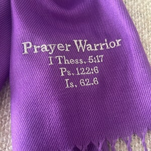 May include: A vibrant purple scarf with a fringed edge. The scarf features the embroidered text "Prayer Warrior" along with Bible verses I Thess. 5:17, Ps. 122:6, and Is. 62:6 in white thread. The scarf is made of a soft, flowing fabric.