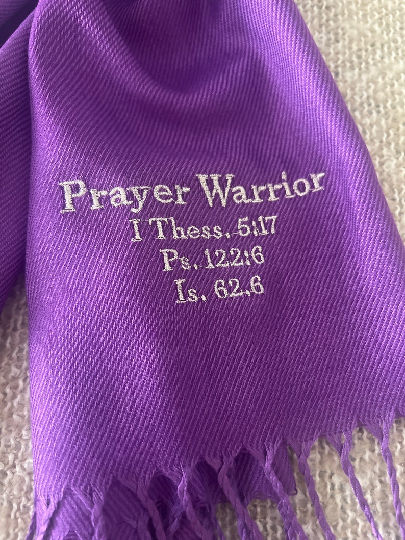 May include: A vibrant purple scarf with a fringed edge. The scarf features the embroidered text "Prayer Warrior" along with Bible verses I Thess. 5:17, Ps. 122:6, and Is. 62:6 in white thread. The scarf is made of a soft, flowing fabric.