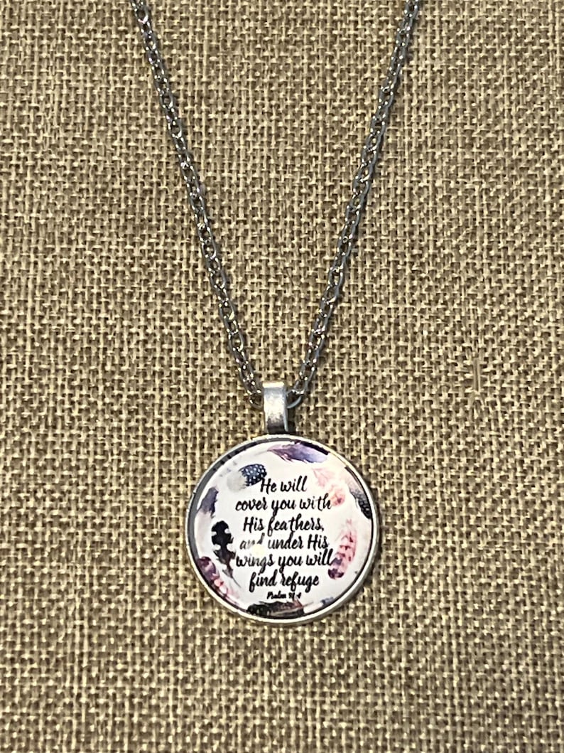 Puede incluir: Un collar de cadena de plata con un colgante redondo que presenta un dise&ntilde;o floral de estilo acuarela y el texto "He will cover you with His feathers, and under His wings you will find refuge. Psalm 91:4".