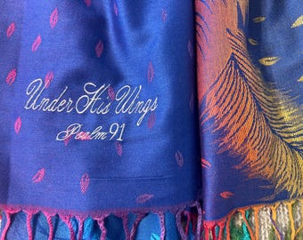 May include: A blue scarf with embroidered text reading "Under His Wings Psalm 91." The scarf features a feather design in shades of orange, yellow, and blue. Pink and purple tassels adorn the bottom edge, adding a decorative touch.