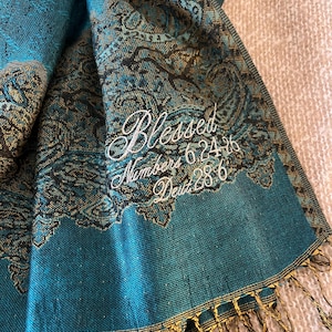 May include: Teal and gold paisley patterned scarf with the word "Blessed" embroidered in white script. The scarf also features the text "Numbers 6.24-26 Deut 28.6" and has a gold fringe.