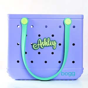 Op de afbeelding: Een lavendelkleurige draagtas met een groene handgreep en de naam "Ashley" in groene glitter. De tas heeft een geperforeerd ontwerp en het woord "bogg" in lichtblauw aan de onderkant.