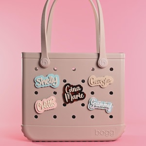 May include: A light pink tote bag with two handles and a perforated body featuring five decorative name tags. The tags display the names Shelly, Gina Marie, Colette, Georgia, and Grammy in various colors and patterns, attached to the bag holes.