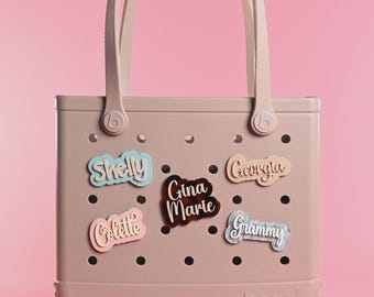 Può includere: Una borsa tote rosa chiaro con due manici e un corpo traforato con cinque targhette decorative con nomi. Le targhette riportano i nomi Shelly, Gina Marie, Colette, Georgia e Grammy in vari colori e motivi, fissate nei fori della borsa.