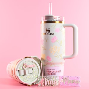 May include: White floral Stanley Quencher H2.0 tumbler with a handle and straw, alongside a smaller tumbler and personalized name tags. The tumblers feature pink and gold accents, with the text "THE FLOWSTATE QUENCHER H2.0 TUMBLER".
