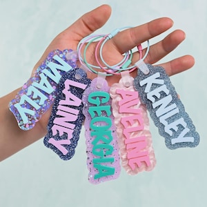 K&ouml;nnte beinhalten: F&uuml;nf rechteckige Kofferanh&auml;nger aus Acryl mit gewelltem Rand, jeweils mit einem erhabenen Namen in einer anderen Farbe: MAELY, LAINEY, GEORGIA, AVELINE und KENLEY. Jeder Anh&auml;nger hat eine glitzernde Oberfl&auml;che und eine farbige Metallschlaufe.