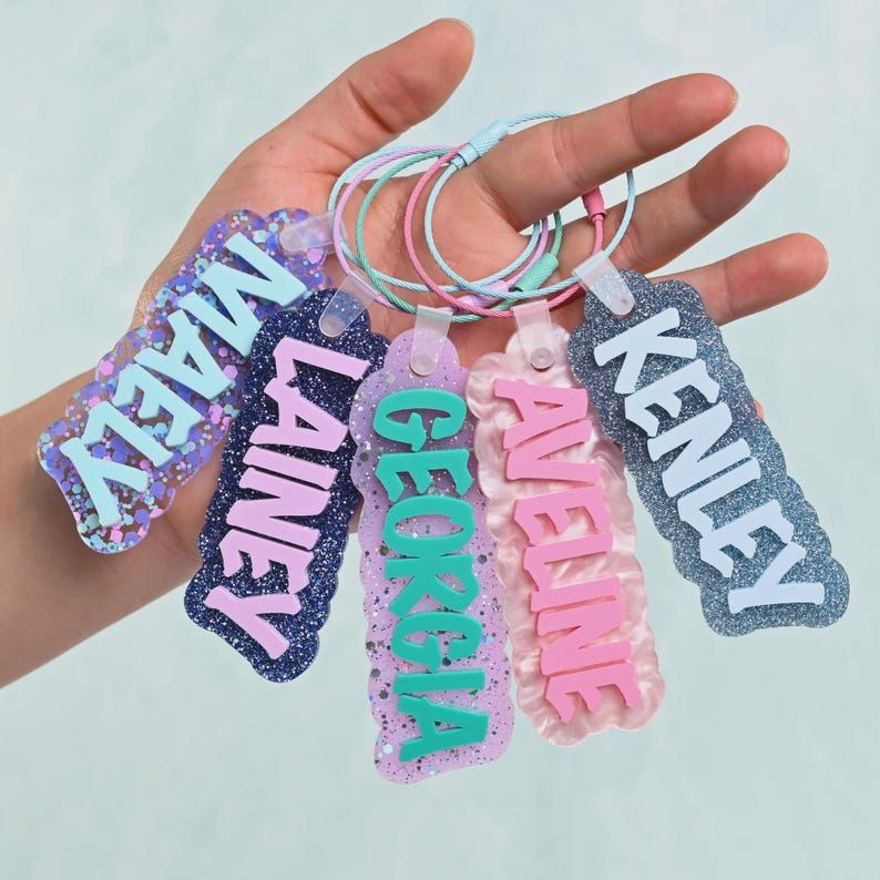 K&ouml;nnte beinhalten: F&uuml;nf rechteckige Kofferanh&auml;nger aus Acryl mit gewelltem Rand, jeweils mit einem erhabenen Namen in einer anderen Farbe: MAELY, LAINEY, GEORGIA, AVELINE und KENLEY. Jeder Anh&auml;nger hat eine glitzernde Oberfl&auml;che und eine farbige Metallschlaufe.