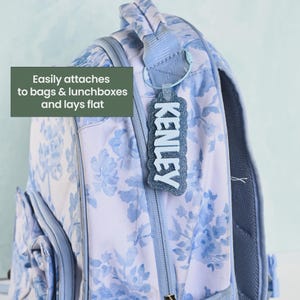 K&ouml;nnte beinhalten: Ein hellblauer Rucksack mit Blumenmuster und einem glitzernden dunkelblauen Gep&auml;ckanh&auml;nger am Griff. Der Anh&auml;nger zeigt den Namen KENLEY in wei&szlig;en Buchstaben. Der Text auf dem Bild lautet: Easily attaches to bags & lunchboxes and lays flat.