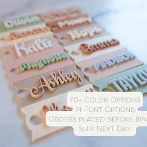 May include: A close-up of personalized name tags in various colors and fonts. The tags are made of acrylic and have a hole for hanging. The text on the tags includes the names "Olivia", "Hope", "Emma", "Madison", "Taylor", "Katie", "Ashley", and "Magnolia". The text "70+ COLOR OPTIONS 14 FONT OPTIONS ORDERS PLACED BEFORE 8PM ET SHIP NEXT DAY" is also visible in the image.