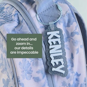 K&ouml;nnte beinhalten: Ein hellblauer Rucksack mit Blumenmuster hat einen glitzernden blauen Kofferanh&auml;nger am Griff. Das Schild zeigt den Namen KENLEY in wei&szlig;er, erhabener Schrift. Ein gr&uuml;nes Feld mit dem Text Go ahead and zoom in... our details are impeccable ist auf der linken Seite des Bildes eingeblendet.
