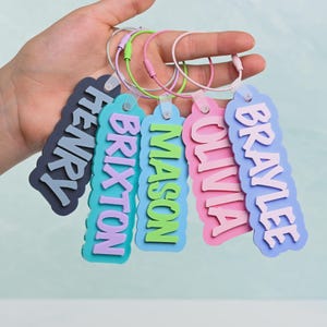 Op de afbeelding: Vijf rechthoekige bagagelabels met geschulpte randen, elk voorzien van een verhoogde naam in een contrasterende kleur. De labels zijn gelabeld met Henry, Brixton, Mason, Olivia en Braylee en bevestigd aan individuele kleurrijke metalen ringen.
