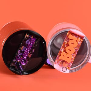 May include: Two insulated tumblers with lids. One is black with a purple glitter name, "Samantha". The other is pink with orange glitter and the word "Boo!". Both are on an orange background.