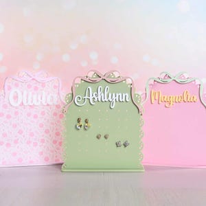 May include: Three personalized earring holders in pastel colors. The center holder is light green with the name "Ashlynn" in white script. The other two are pink, with the names "Olivia" and "Magnolia". Each has a decorative bow and scalloped edges.