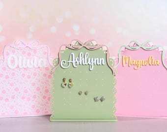 May include: Three arched earring display stands with scalloped edges and bow-shaped tops. The left stand is pink with floral patterns and the name Olivia. The centre stand is sage green with the name Ashlynn. The right stand is solid pink with the name Magnolia.