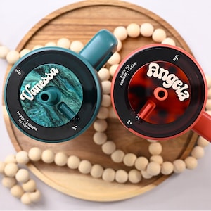 May include: Two personalized drink lids, one teal and one coral, each with a name in white script. The teal lid has a teal and green marbled interior, while the coral lid has a tortoiseshell pattern. Both lids have black rims and handles.