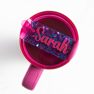 Könnte beinhalten: Eine rosa Reise-Tasse mit Henkel und Deckel. Die Tasse hat einen glitzernden blau-rosa Aufkleber mit dem Namen "Sarah" darauf.