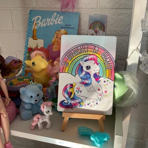 May include: A colorful My Little Pony illustration on a small easel. The pony is pink and blue with a rainbow mane and tail. The text on the illustration reads "So Apparently I'm Dramatic".