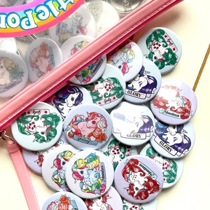 May include: A collection of colourful My Little Pony buttons featuring different characters and names such as "Galaxy", "Moondancer", and "Gusty". The buttons are in a clear plastic bag with a pink zipper.