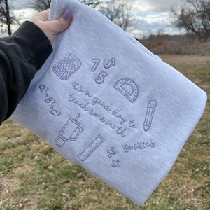 May include: A light gray sweatshirt embroidered with math-related designs. The design includes a calculator, protractor, pencil, and the phrase "it's a good day to teach some math." Also includes mathematical equations and numbers.